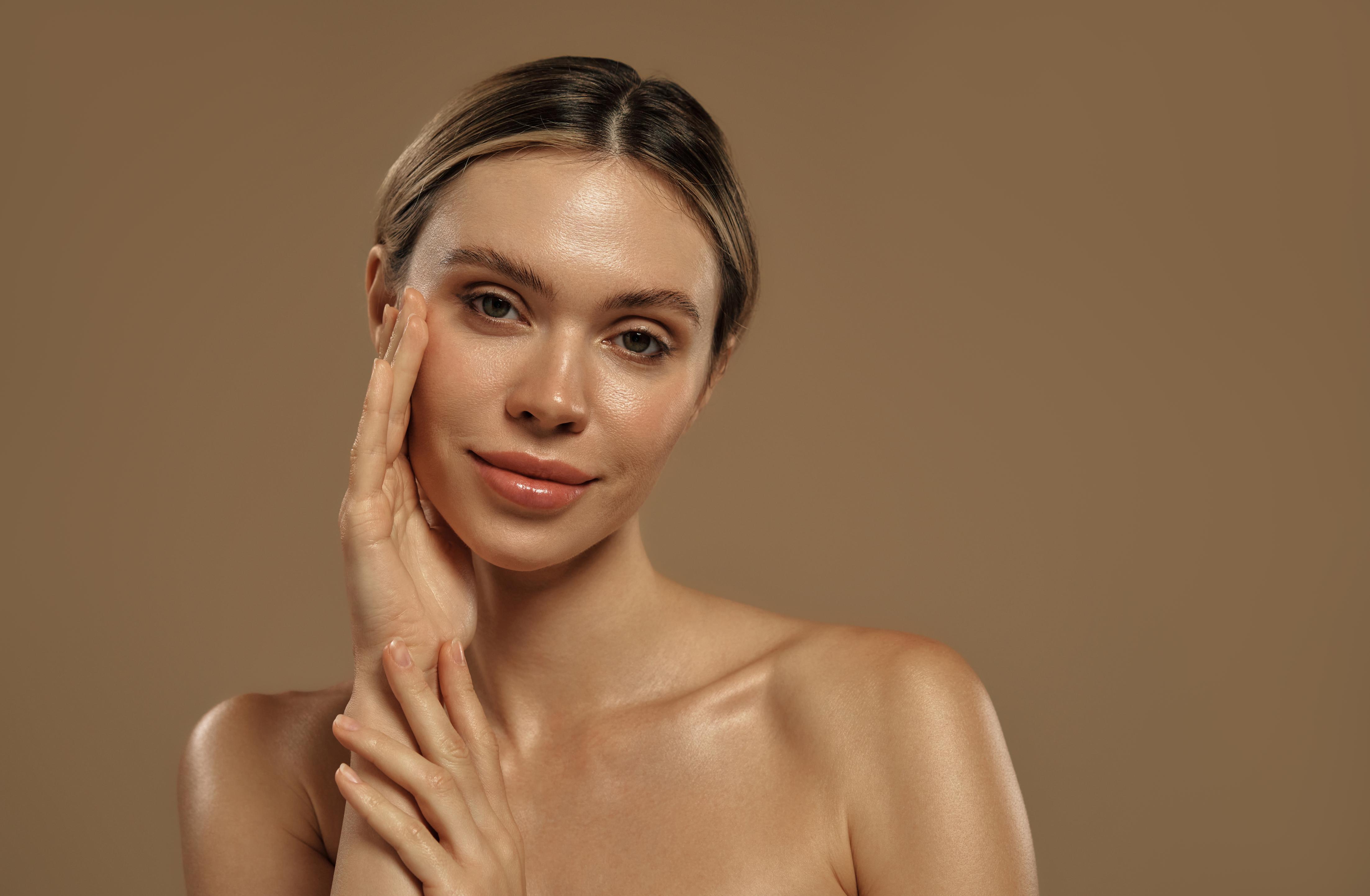 Portrait of a young woman with clear skin gently touching her face against a brown background.