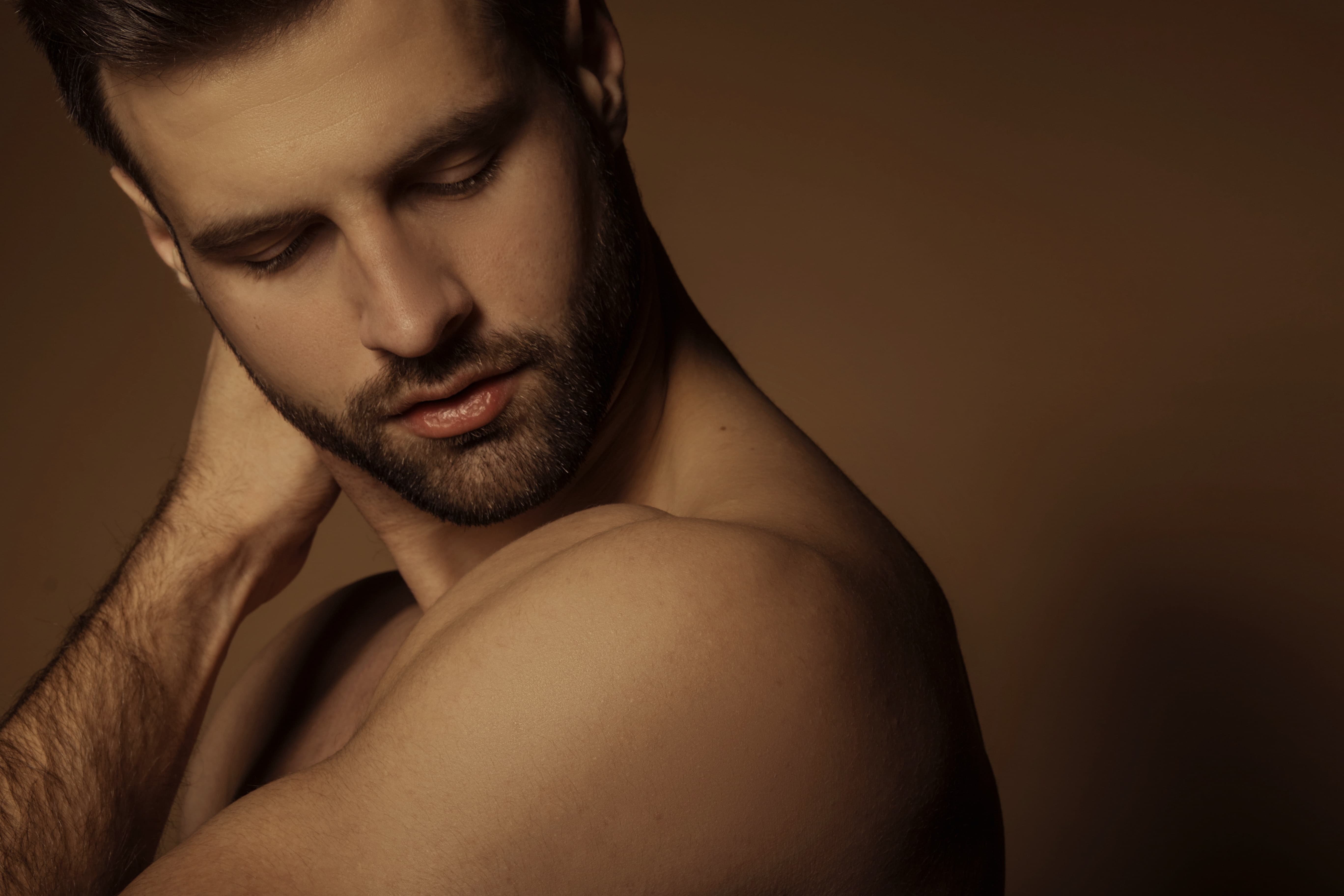 Bearded man with closed eyes touching the back of his neck against a brown background.