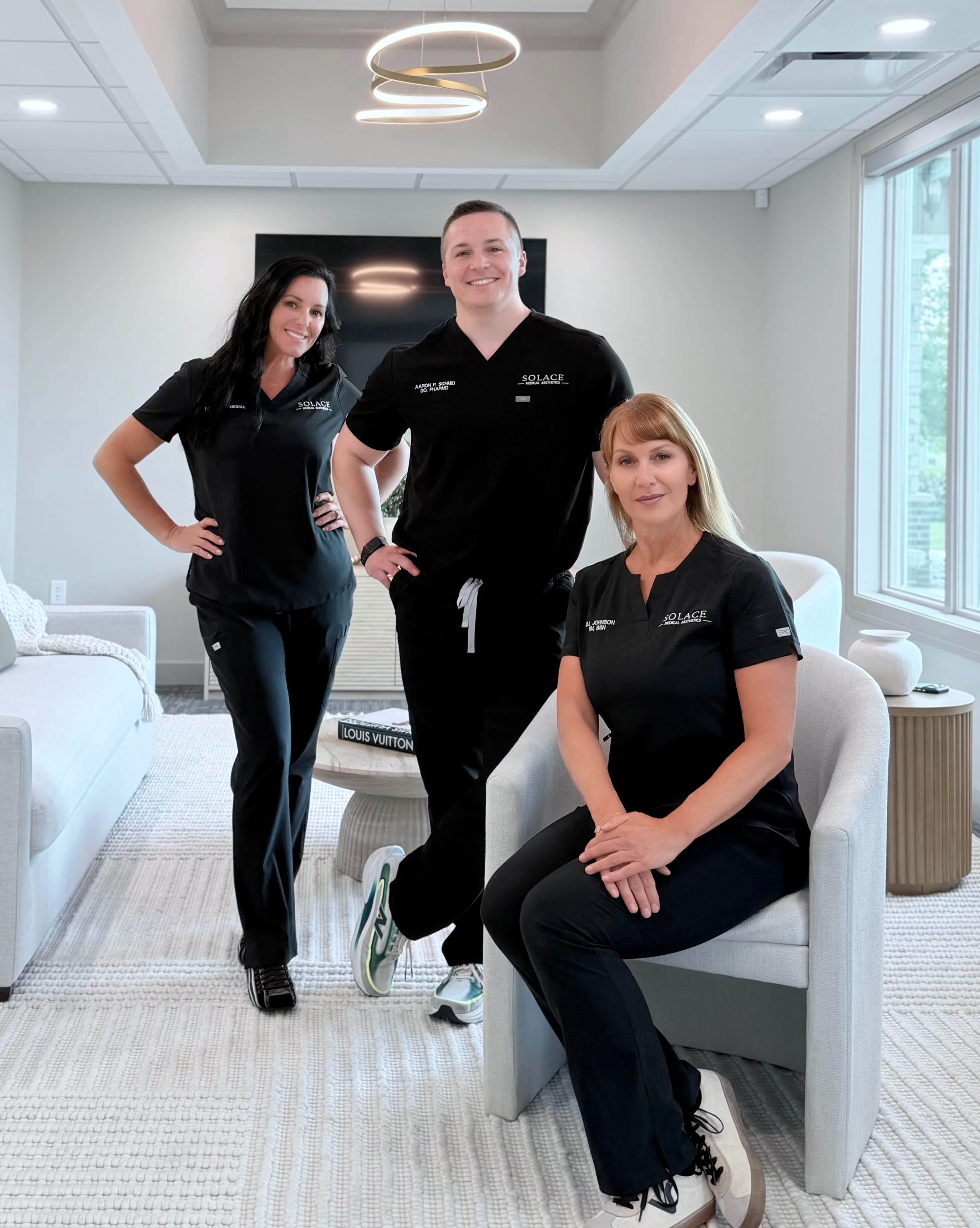 Three medical professionals in black scrubs posing in a modern, bright office with white furniture.