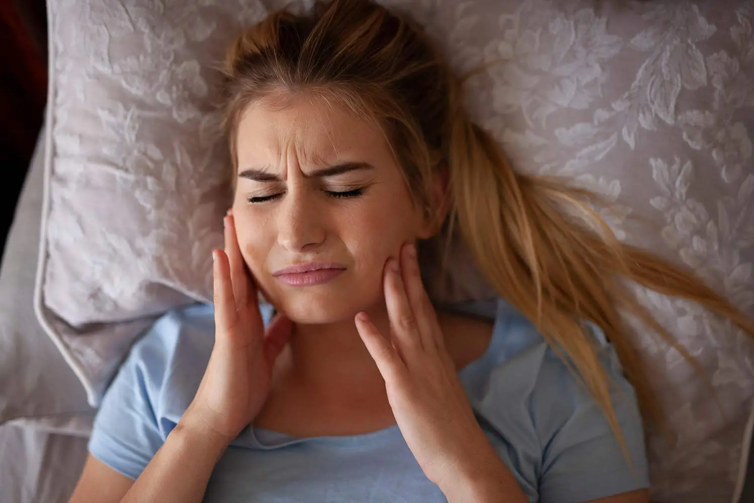 Woman lying on a pillow with eyes closed and hands on her cheeks, showing discomfort or pain.