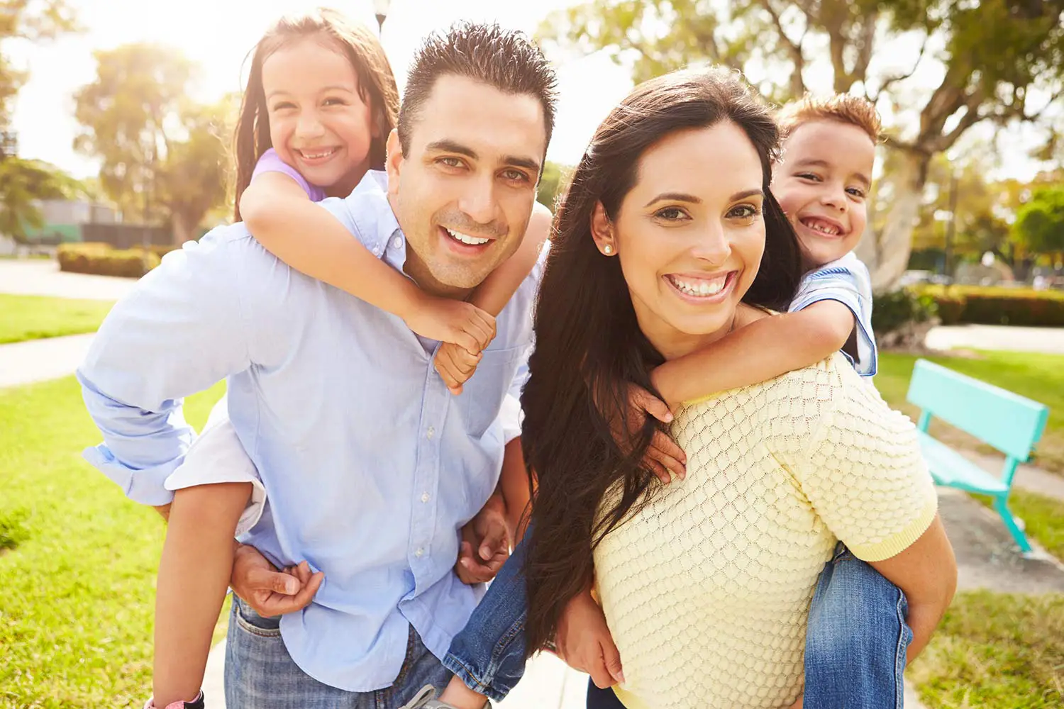Smiling parents giving piggyback rides to their two children outdoors in a sunny park.