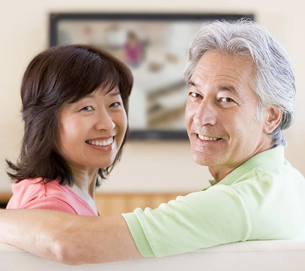 Smiling middle-aged Asian woman and man sitting closely on a sofa, looking at the camera.