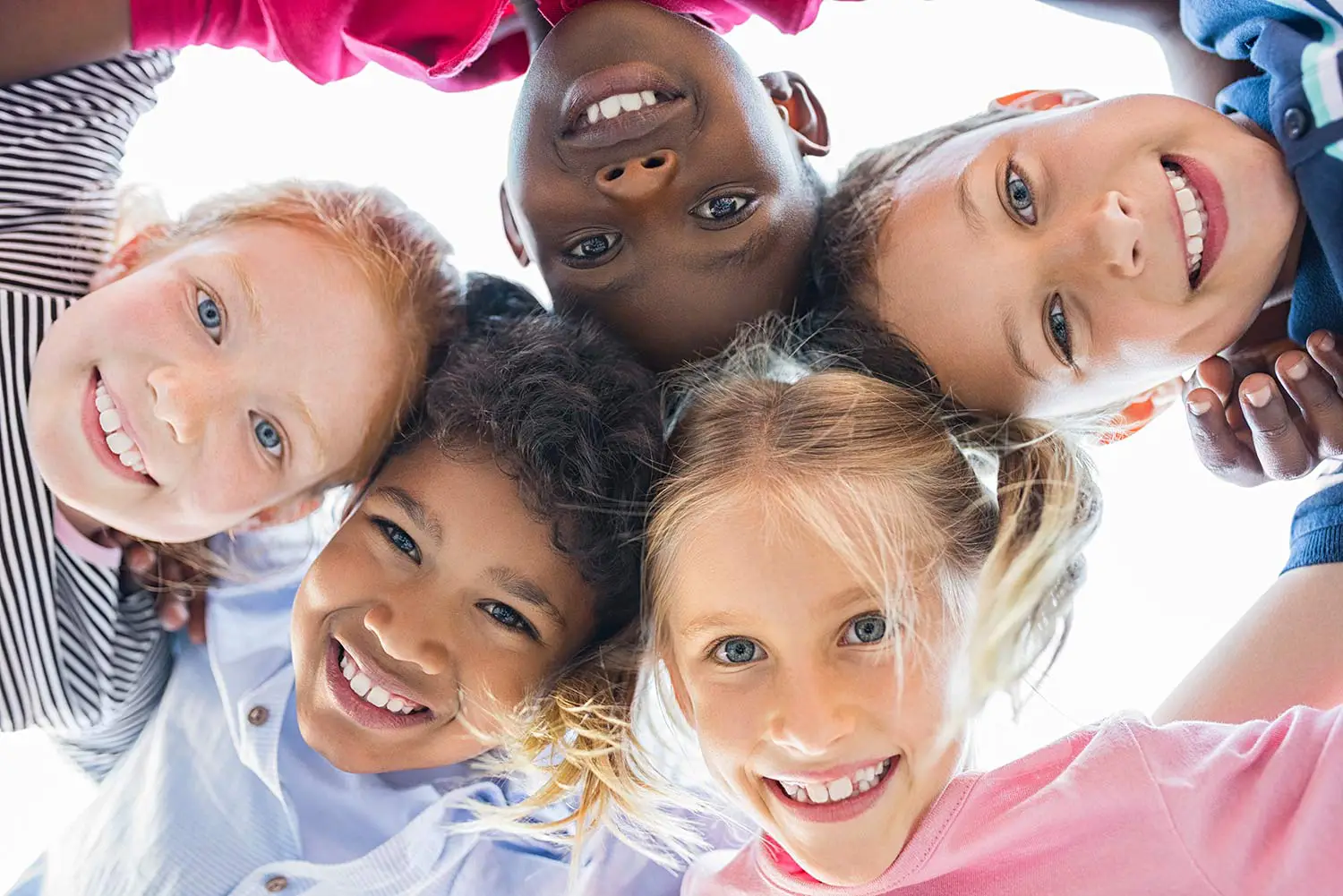 Group of five diverse children smiling and leaning in a circle, looking down at the camera.