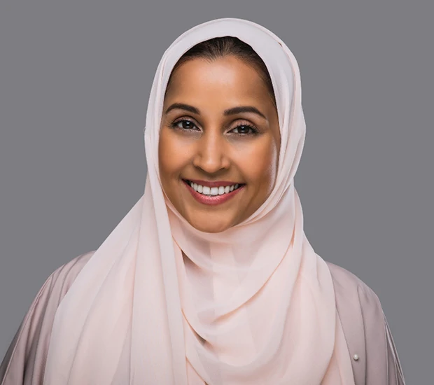 Smiling woman wearing a light pink hijab and matching outfit against a gray background.