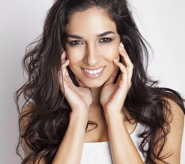 Smiling woman with long dark hair holding her face with both hands.