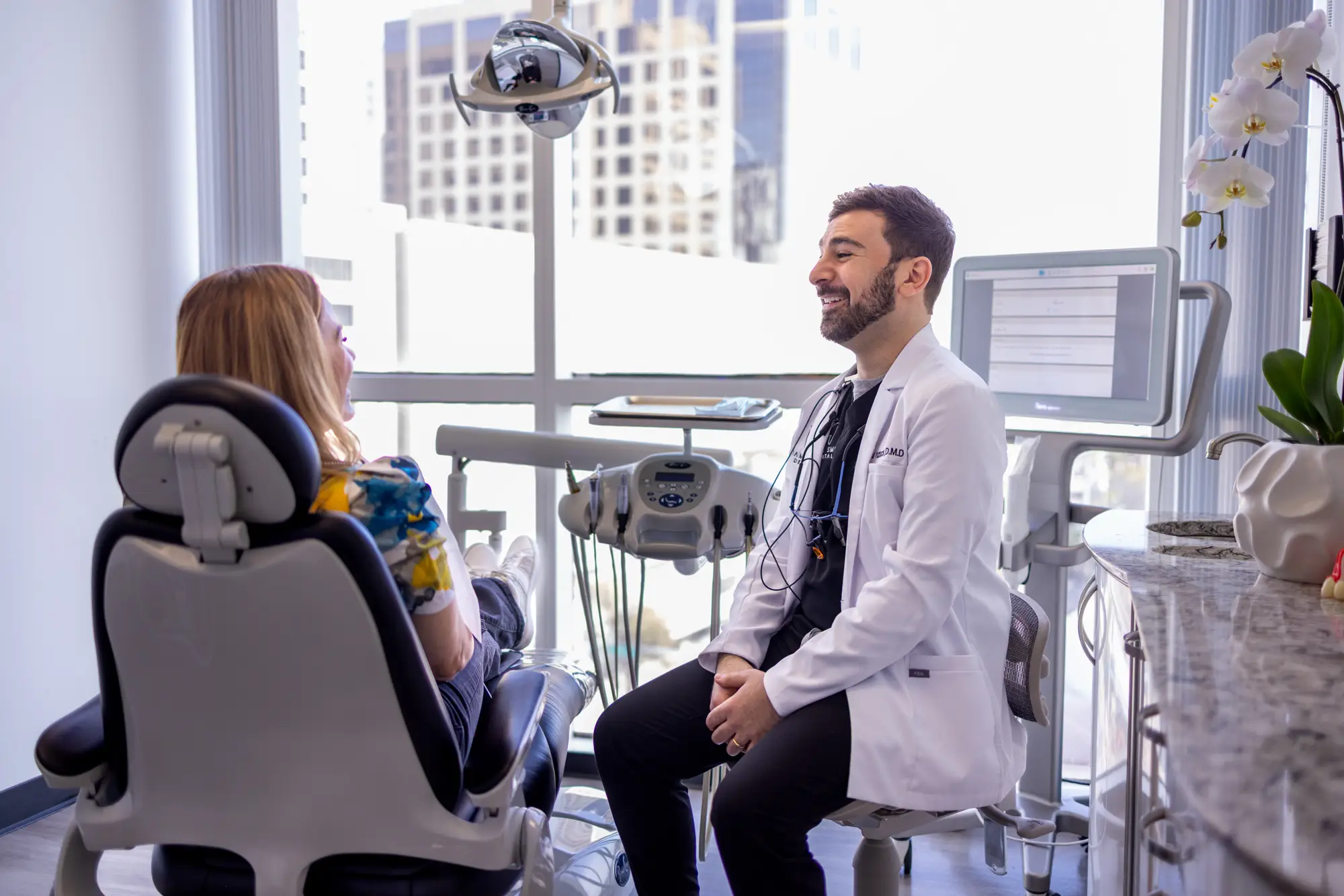 Dentist in a white coat talking and smiling with a female patient seated in a dental chair by a large window.