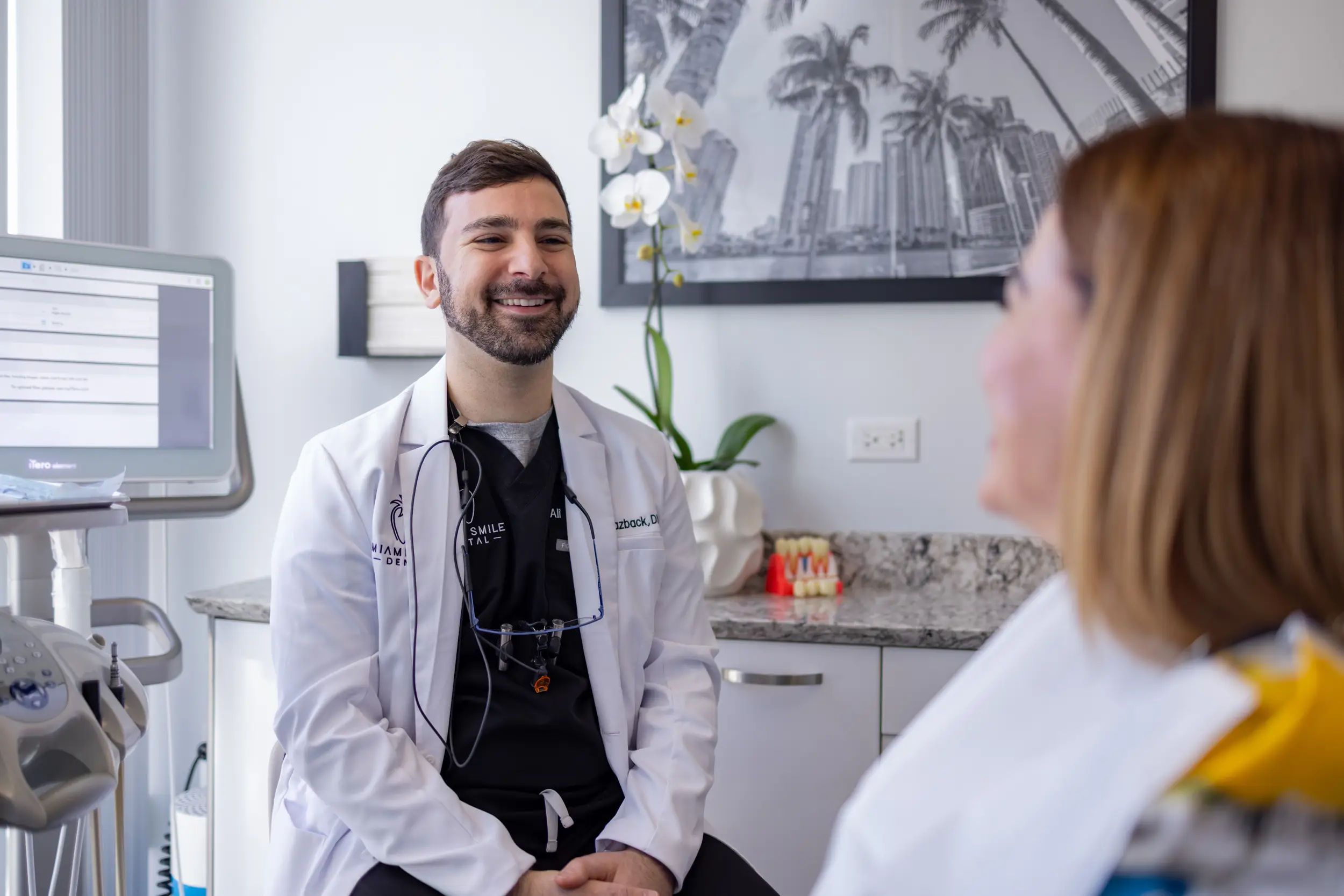 Male dentist in white coat smiling and talking to a female patient in a dental office.