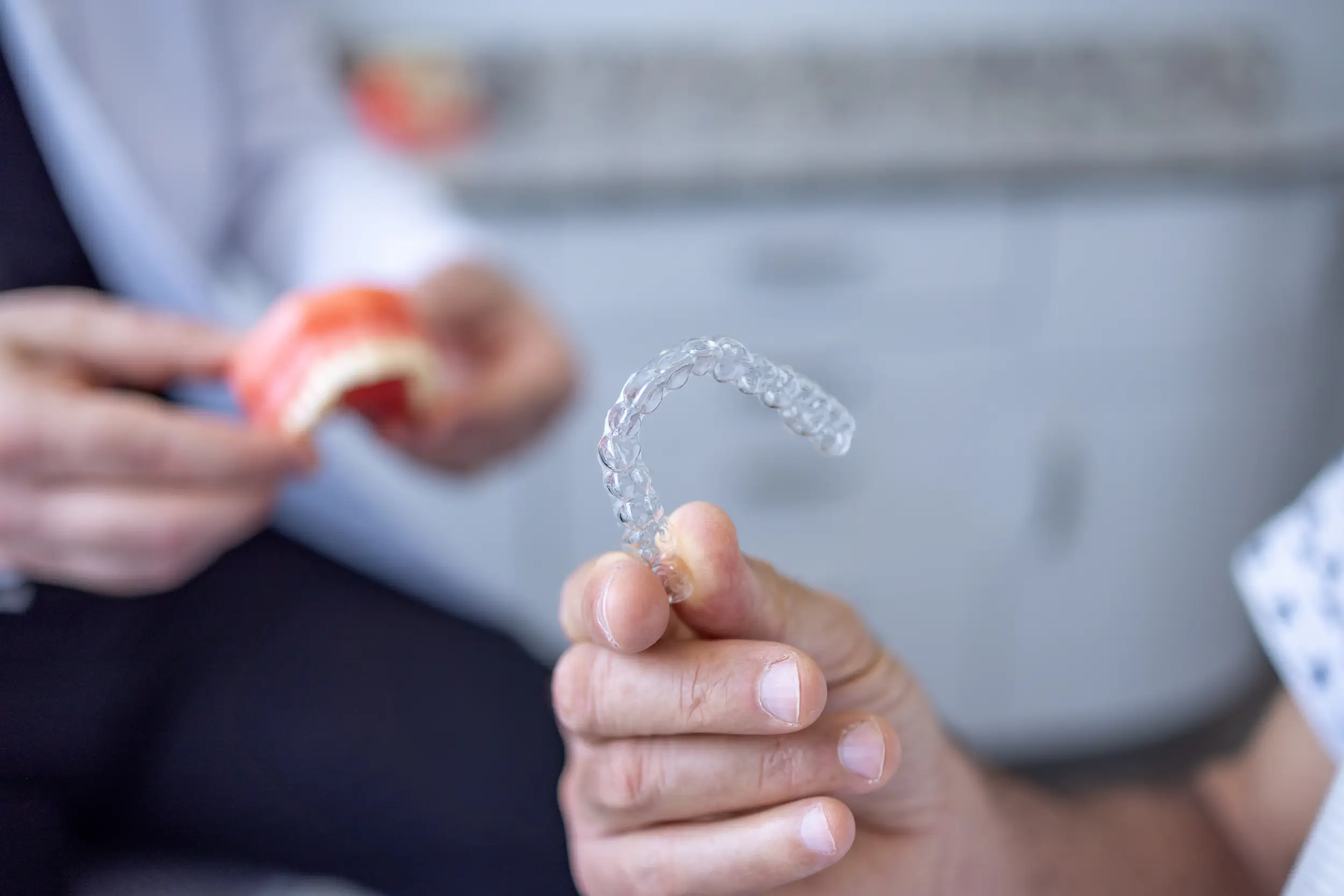 Hand holding a clear dental aligner with a blurred figure holding a dental model in the background.