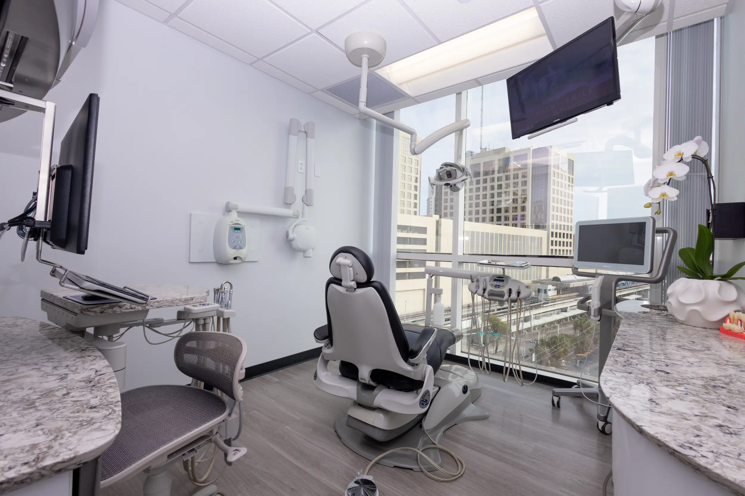 Modern dental clinic room with dental chair facing large windows overlooking city buildings, dental equipment, monitors, and a white orchid on the counter.