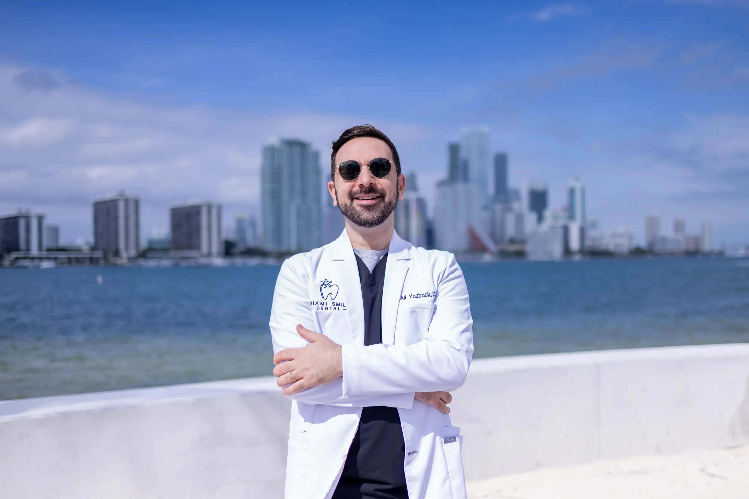 Smiling male dentist in white coat and sunglasses standing with arms crossed by waterfront with city skyline in the background.