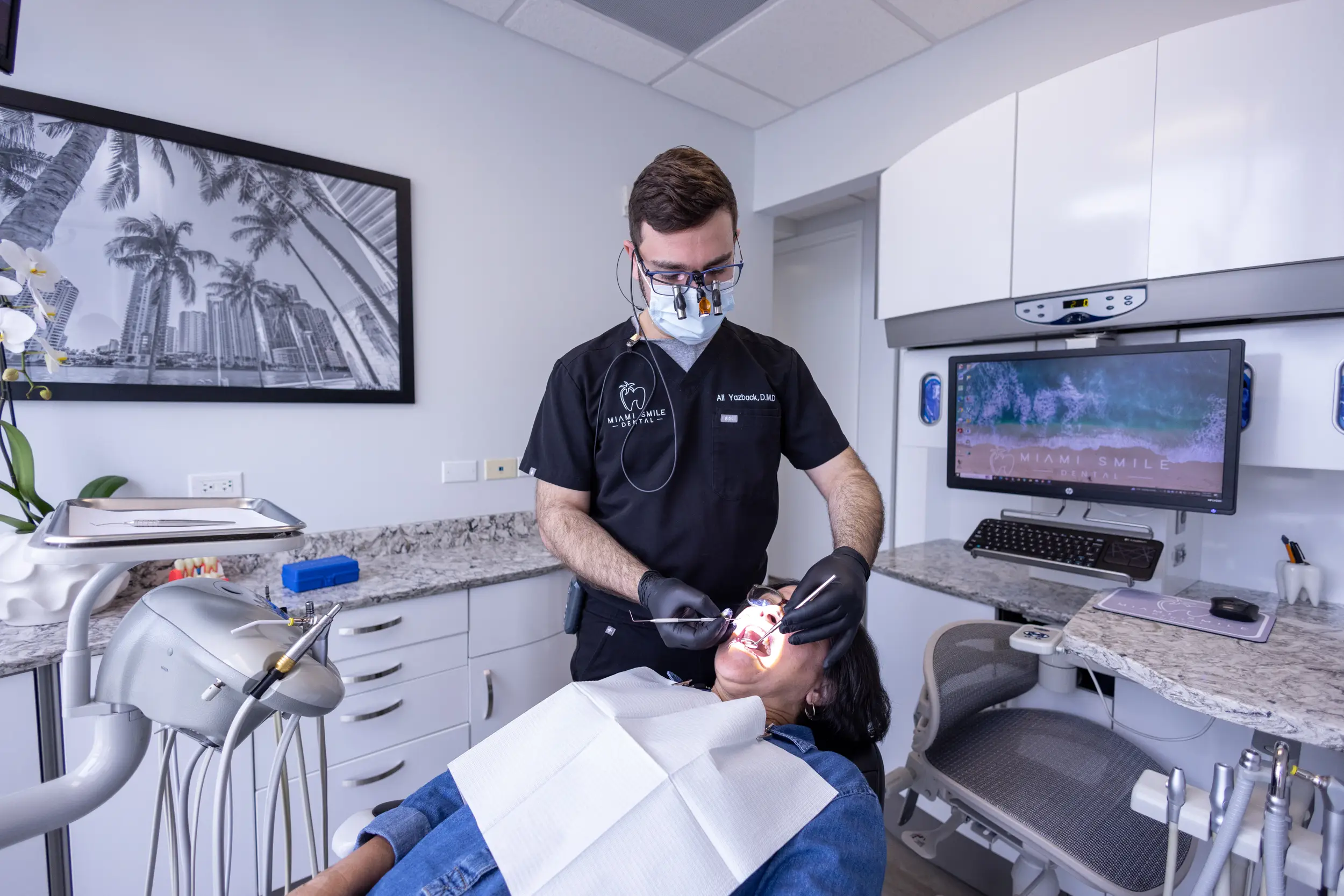 Dentist wearing black scrubs and magnifying glasses examining a patient's mouth in a modern dental clinic.