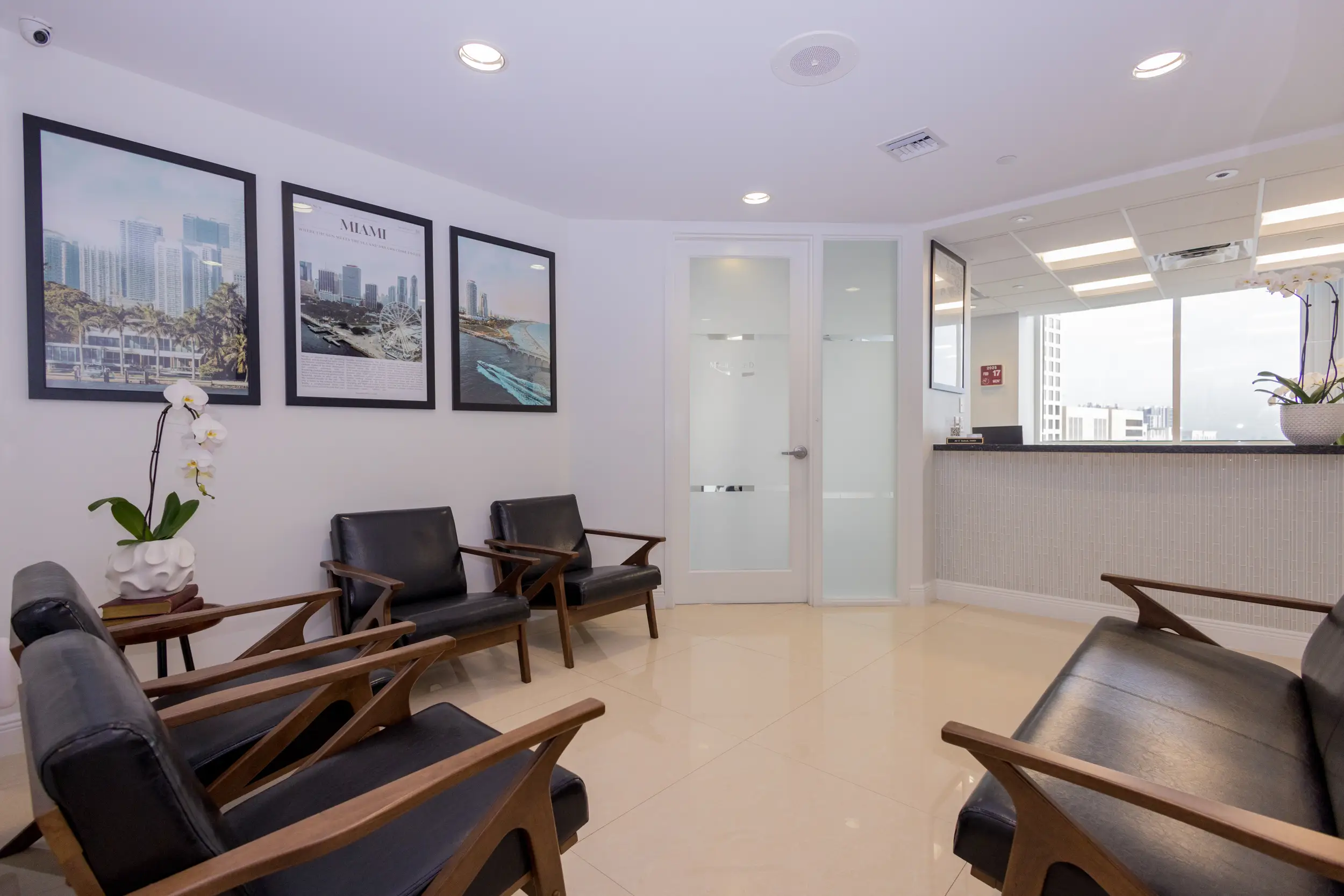 Modern waiting room with black leather chairs, Miami-themed framed photos on the wall, a white orchid on a wooden table, and a reception desk near a window.