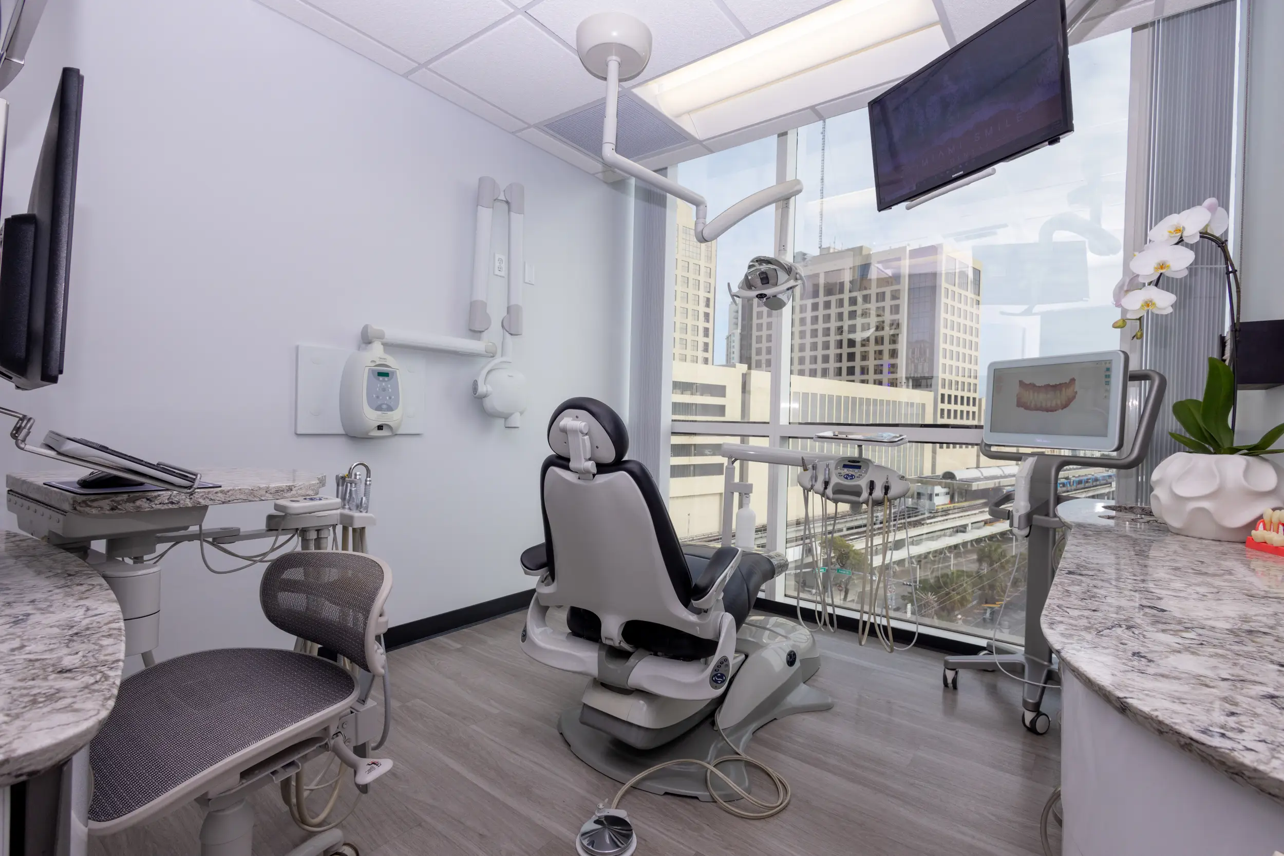 Modern dental office with a dental chair facing large windows showing a cityscape, dental equipment, monitors, and a white orchid on a marble countertop.