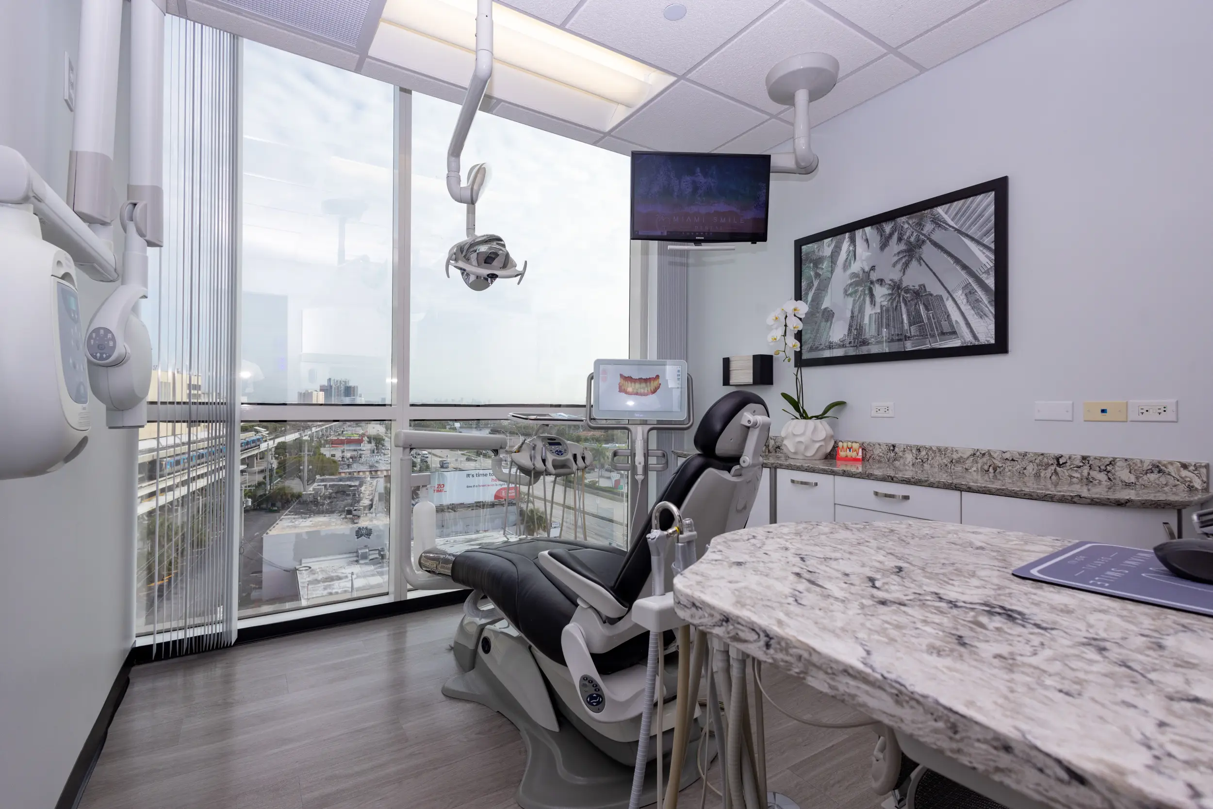 Modern dental office with a black dental chair facing large floor-to-ceiling windows overlooking a cityscape, dental equipment, a TV mounted on the wall, and a framed black and white photo of palm trees and buildings.