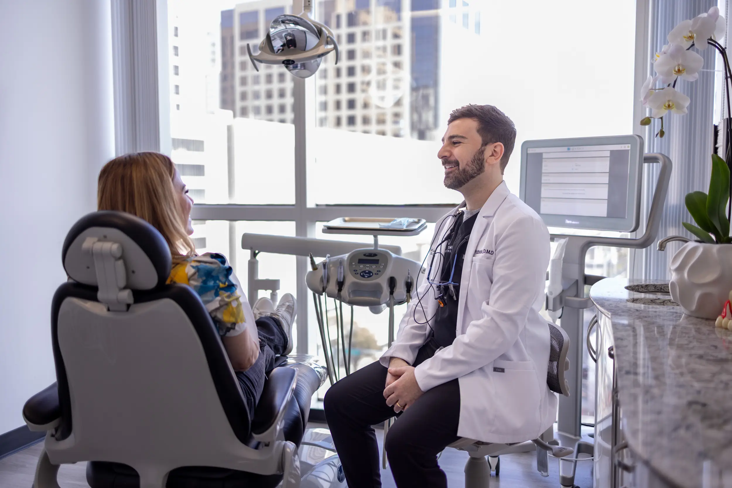 Dentist in white coat talking with a female patient seated in a dental chair in a modern clinic.
