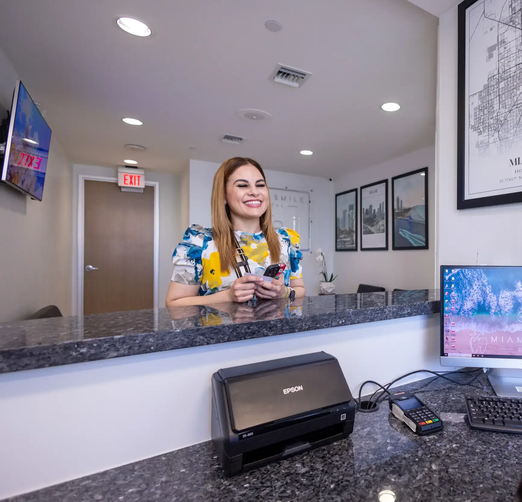 Smiling woman standing behind a reception desk in a modern office with framed cityscape pictures and exit signs.