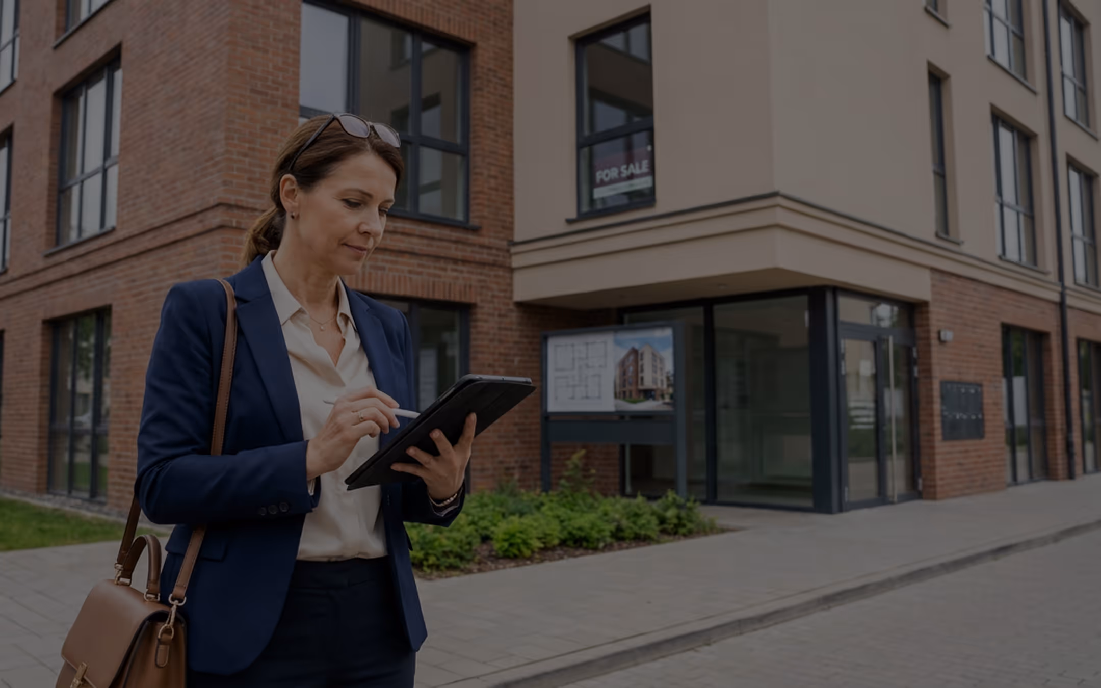 Frau in Anzug nutzt Tablet vor einem modernen Gebäude mit "Zu verkaufen"-Schild.