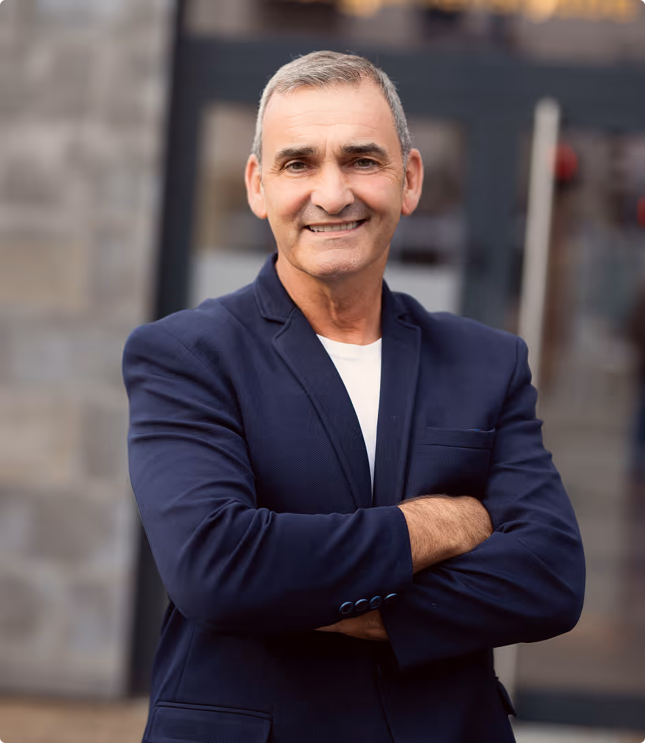 Älterer Mann mit grauen Haaren und blauem Blazer lächelnd mit verschränkten Armen.