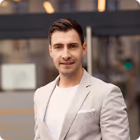 Junger lächelnder Mann in hellgrauem Sakko und weißem T-Shirt vor städtischem Hintergrund.