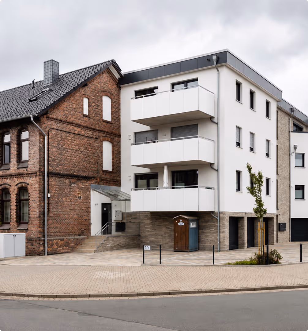 Modernes weißes Mehrfamilienhaus neben einem älteren ziegelroten Gebäude an einer ruhigen Straße.
