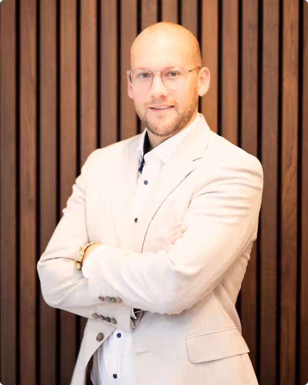 Mann mit Brille und hellbeiger Jacke vor dunkler Holzplatte, Arme verschränkt, lächelnd.
