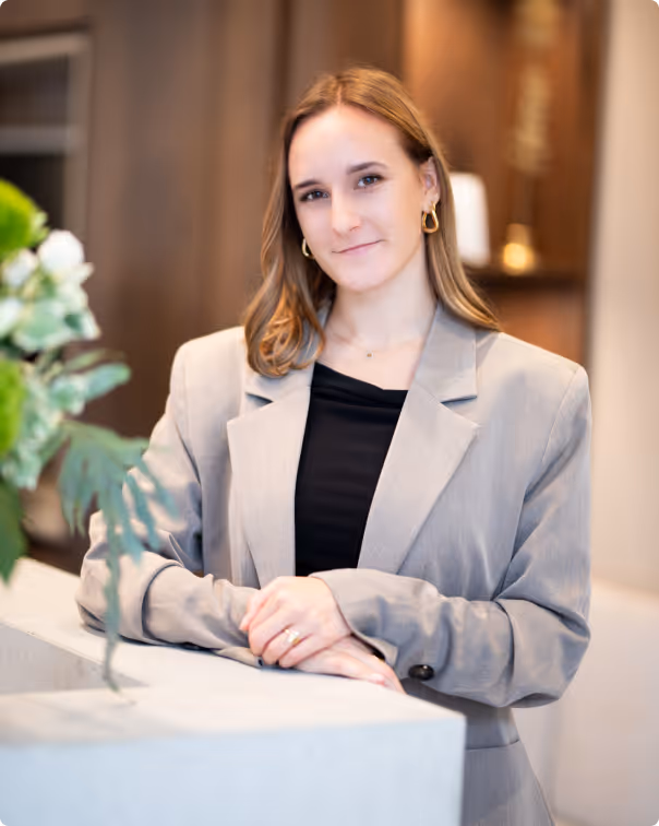 Junge Frau in hellem Blazer und schwarzem Oberteil lehnt an weißer Oberfläche, lächelt.