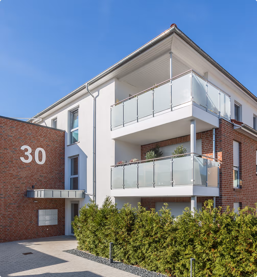 Modernes Wohngebäude mit zwei Balkonen, rotem Backstein und grüner Hecke bei klarem Himmel.