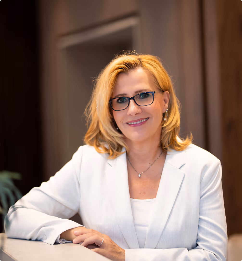 Lächelnde blonde Frau mit Brille in weißem Blazer an einem Tisch im Büro.