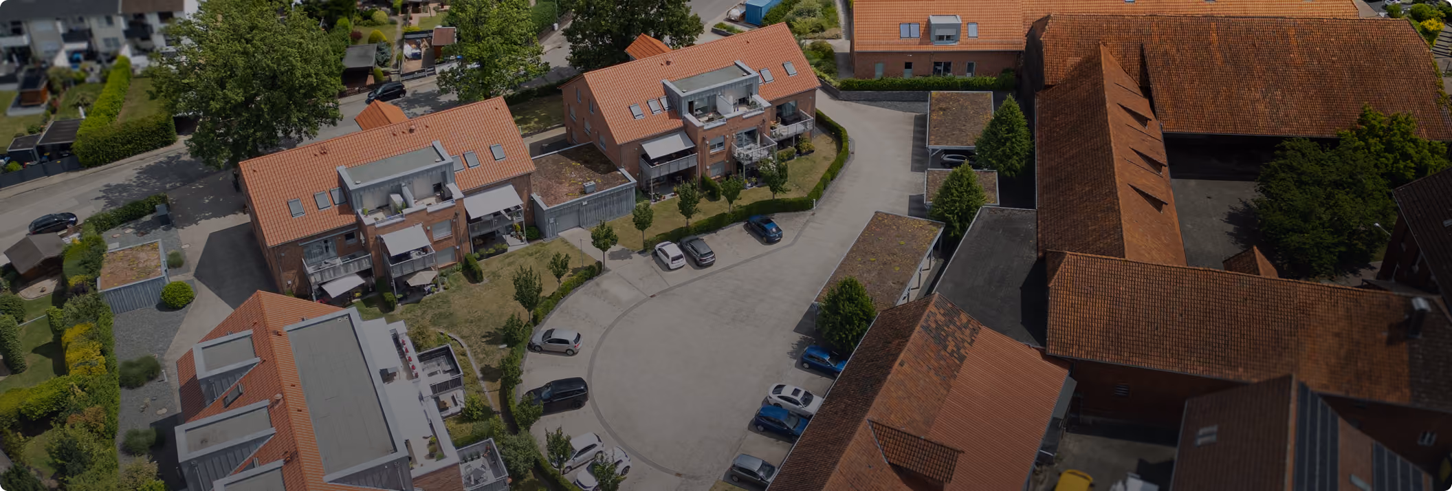 Luftaufnahme eines Wohngebiets mit Parkplätzen, Bäumen und roten Dachziegelhäusern.