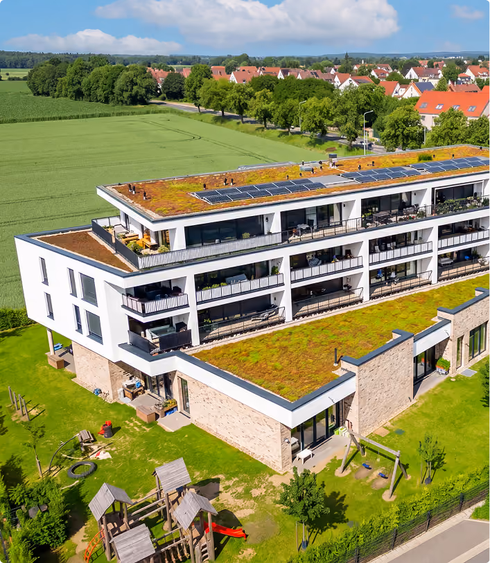 Modernes Wohngebäude mit begrüntem Dach, Solaranlagen, Spielplatz, umgeben von Feldern und Wohnhäusern.