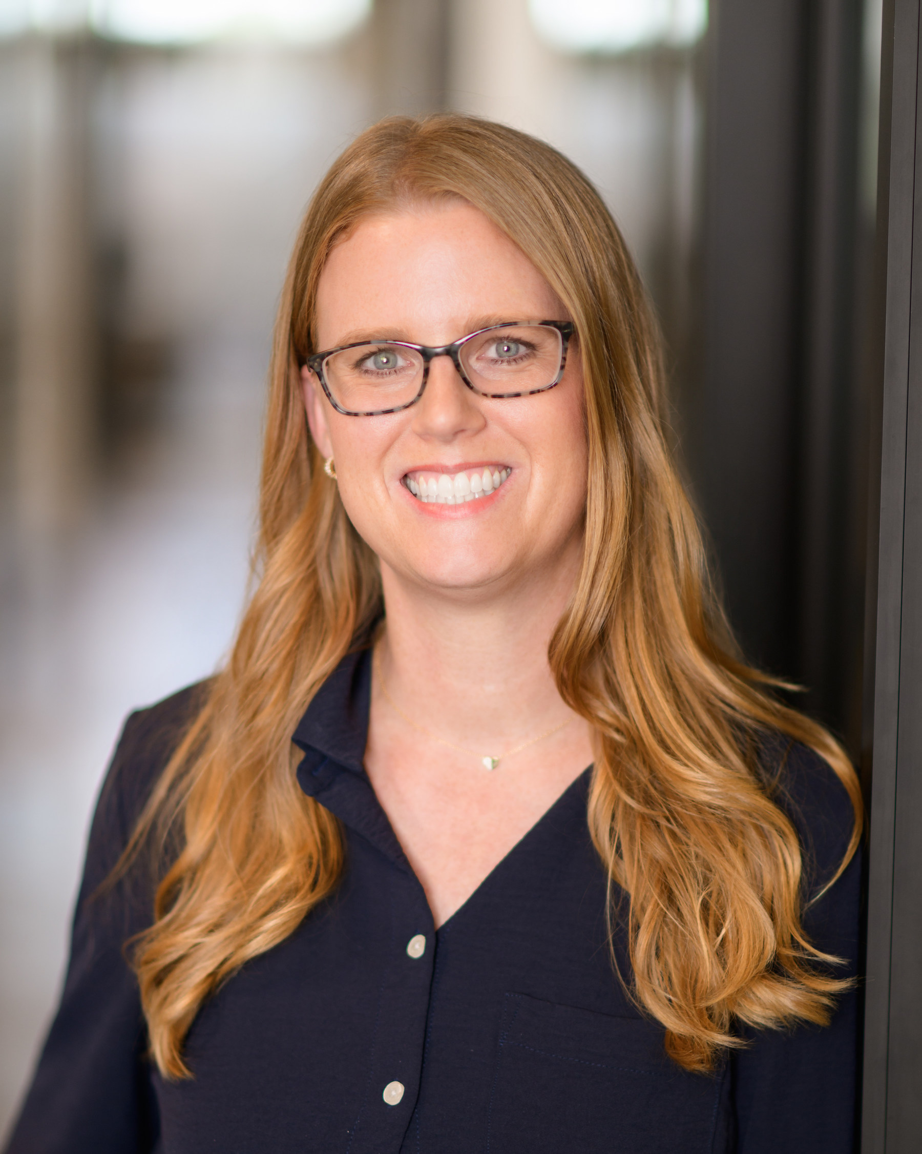 Smiling woman with long blonde hair and glasses wearing a dark button-up shirt indoors.