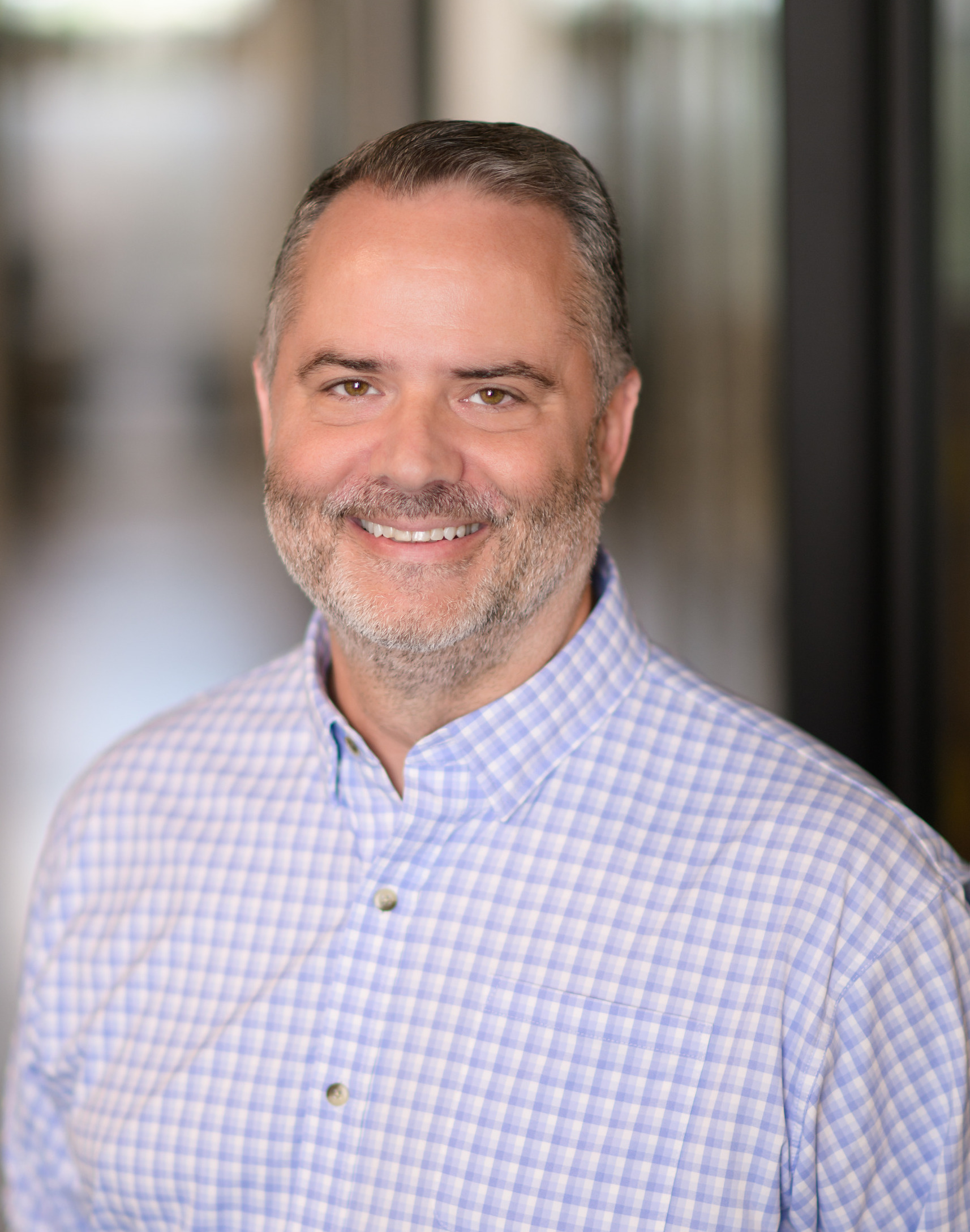 Smiling middle-aged man with short hair and beard wearing a blue checkered button-up shirt.
