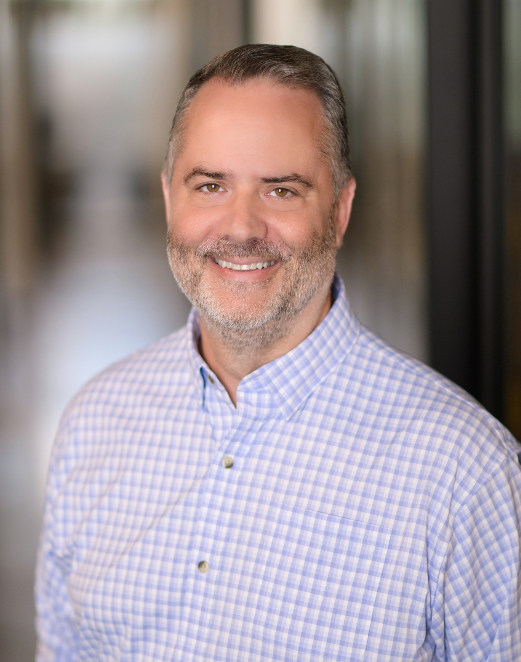 Smiling middle-aged man with short hair and beard wearing a blue checkered button-up shirt.