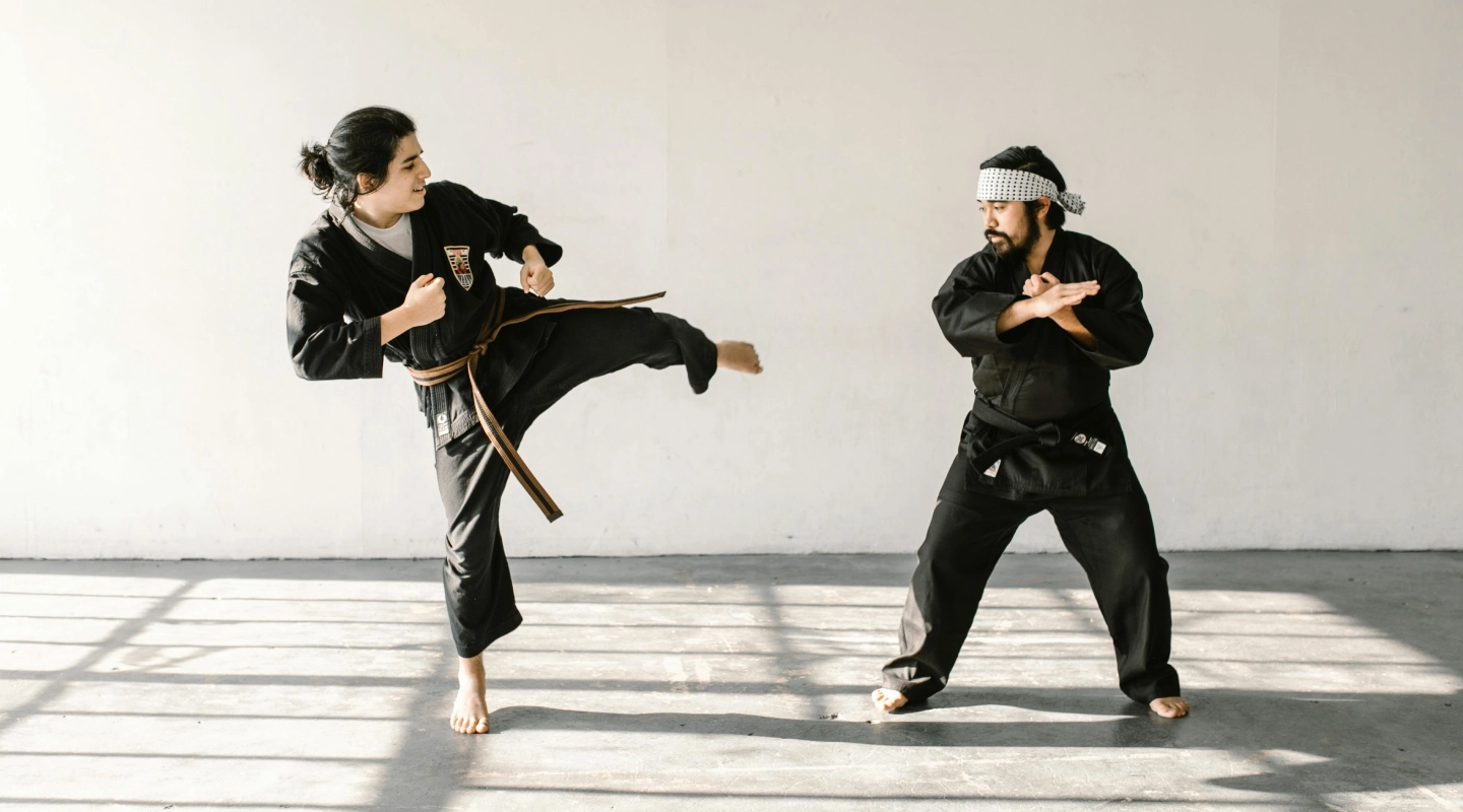 Two martial artists in black uniforms practice sparring in a sunlit room. One executes a kick while the other prepares to block, conveying focus and discipline.