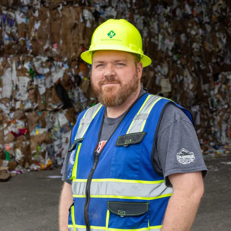 Man in yellow hard hat and blue safety vest stands in front of stacks of recycled cardboard.