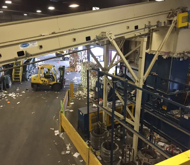 Industrial recycling facility with a yellow forklift, large machinery, and scattered paper debris on floor.