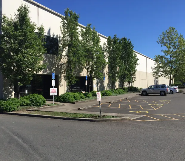 Exterior of a beige commercial building with trees, handicap parking, and a few parked cars under clear blue sky.