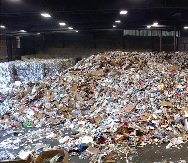 Large piles of mixed recyclable materials and compacted bales inside a recycling facility warehouse.