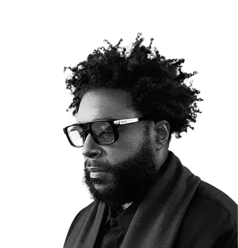 Black man with afro hairstyle, beard, glasses, and dark clothing against a black background.