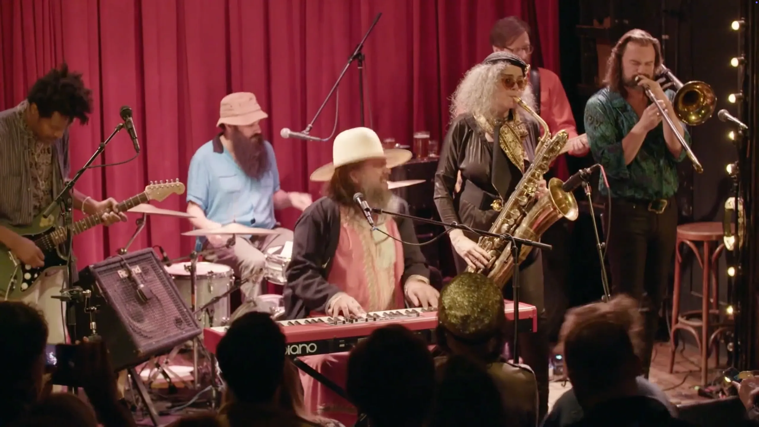 Band performing live with keyboard, saxophone, guitar, drums, trombone, and bass under red curtain.