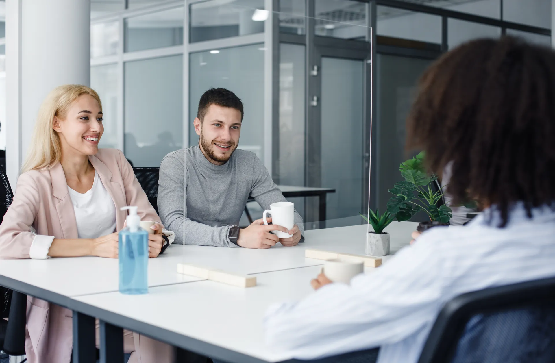 Two people smiling and holding mugs while sitting across a desk from a third person in an office.
