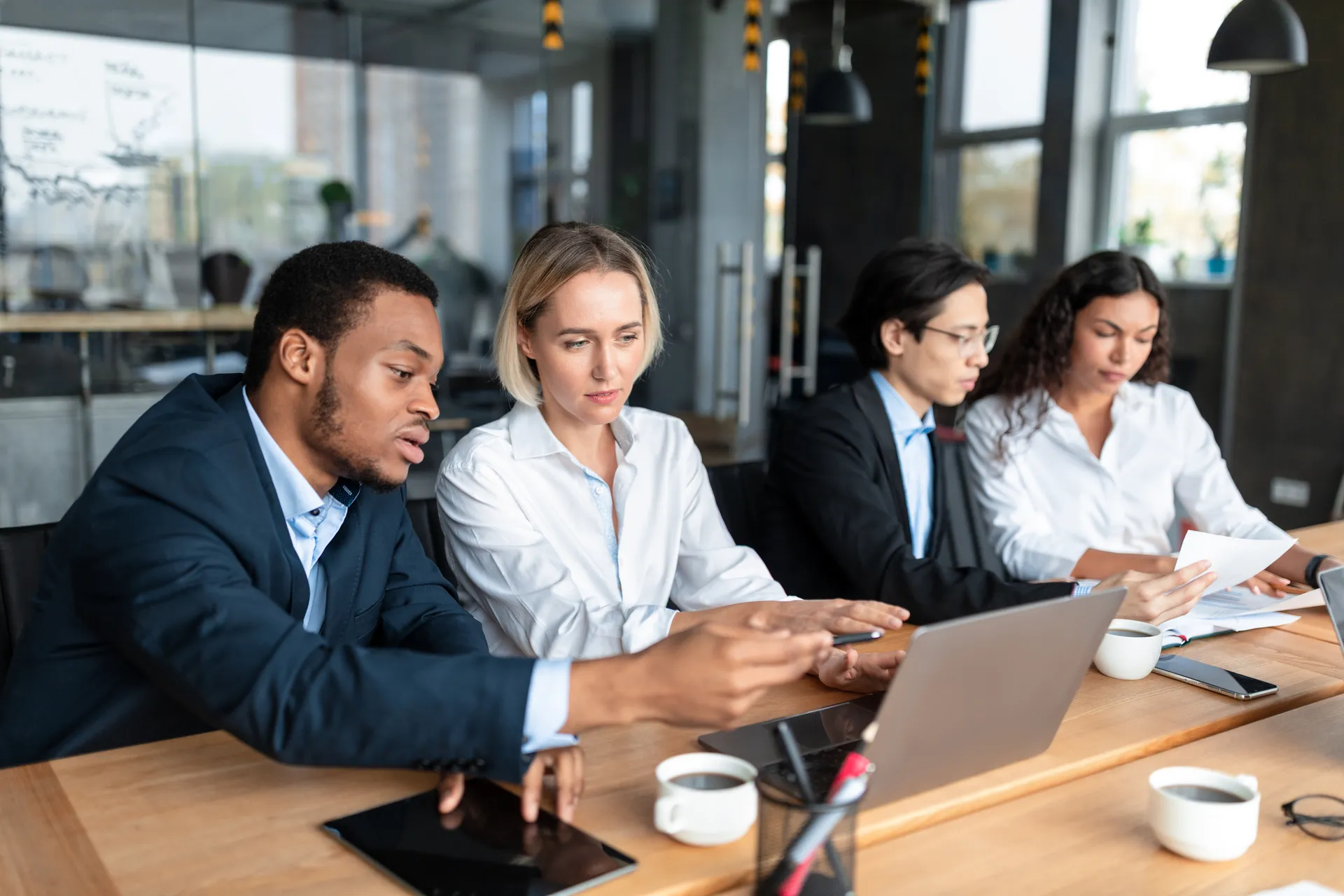 Four diverse coworkers collaborate at a wooden table with laptops, tablets, papers, and coffee cups.