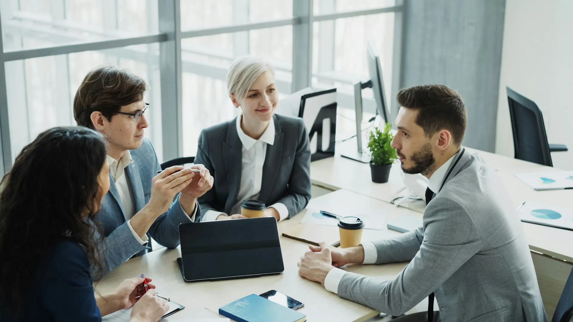 Four colleagues in business attire having a meeting around a table with laptops and coffee cups.