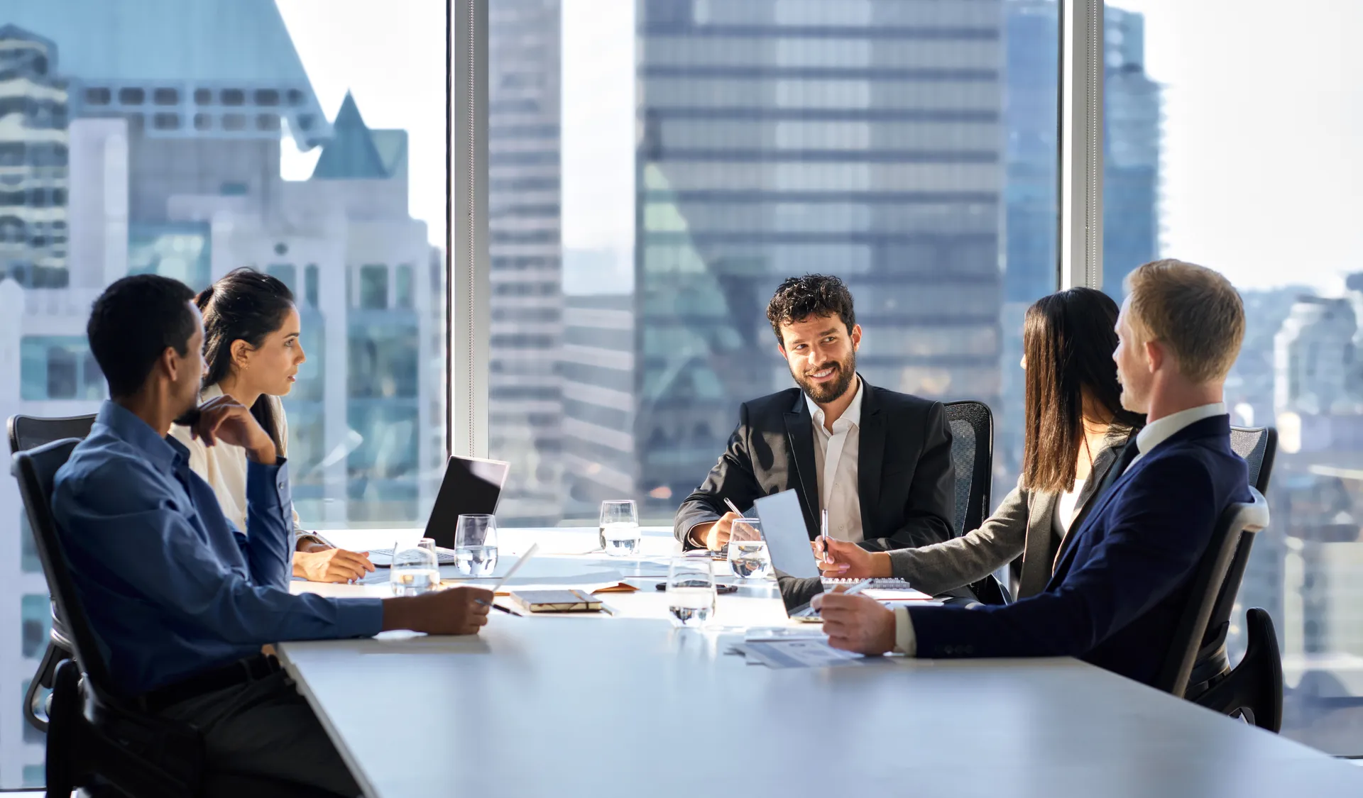 Five businesspeople seated around a conference table in a high-rise office during a meeting.