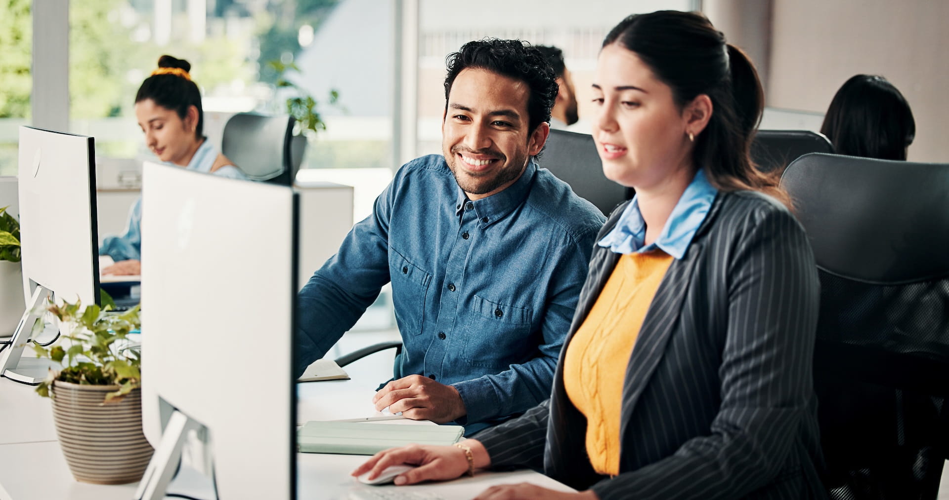 Man and woman collaborating at a computer in a modern office with others working nearby.