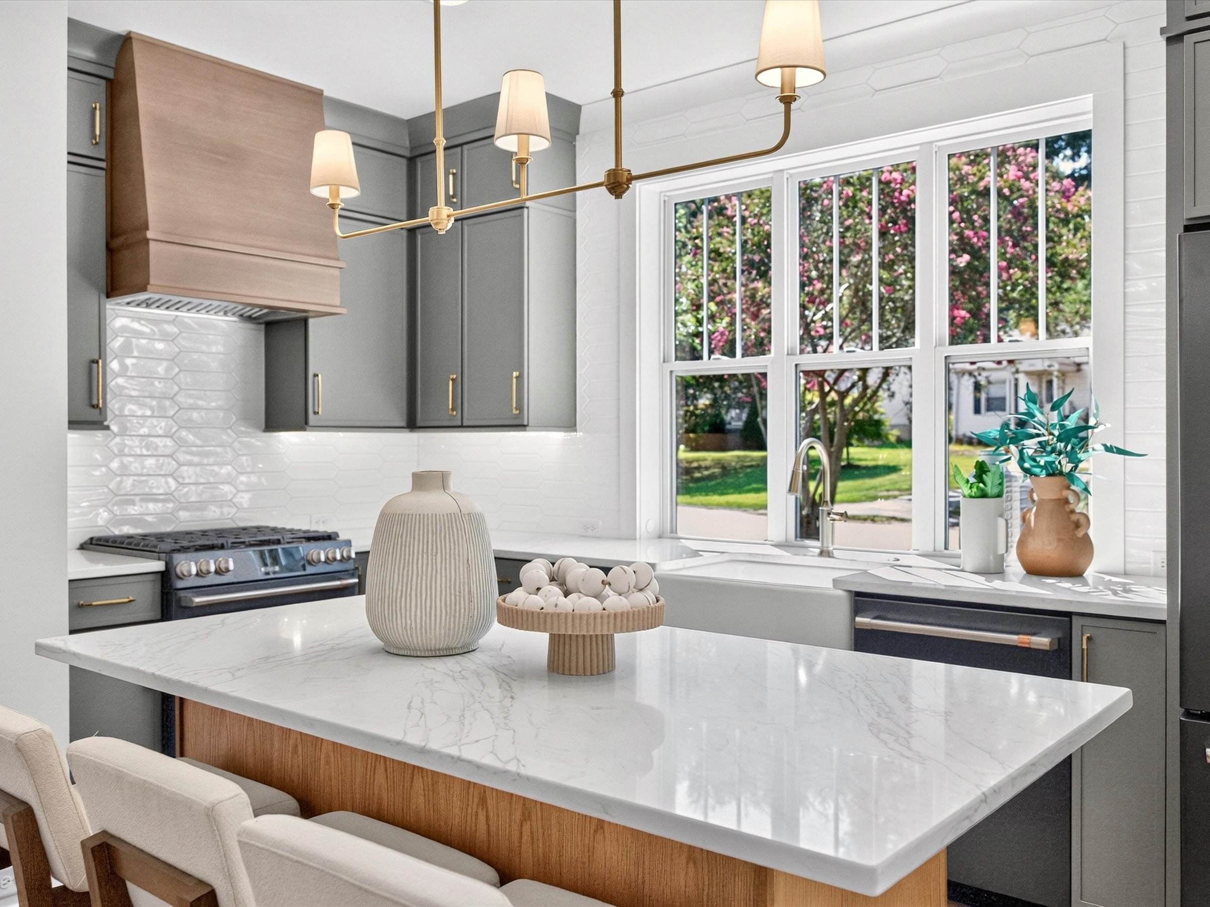 Kitchen island with a white marble top, gray cabinets, a wood range hood, brass pendants, and a large window over the sink