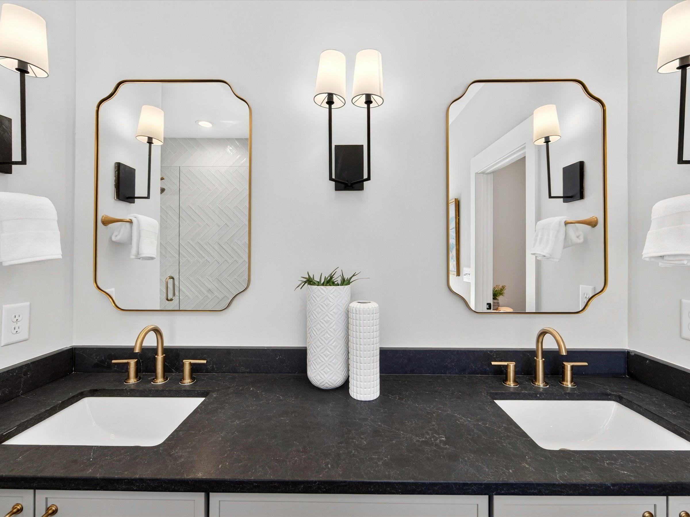 Bathroom vanity with two sinks, a black countertop, brass faucets, arched mirrors, and wall sconces