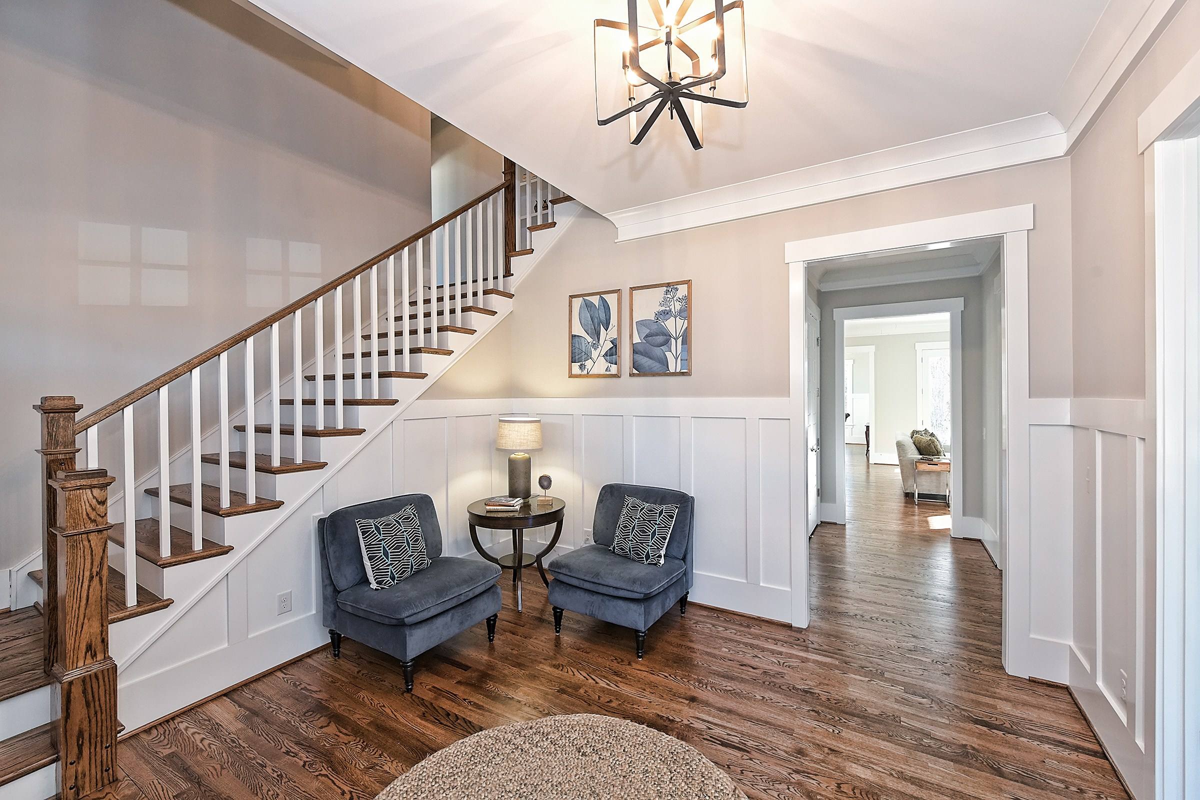 Foyer with a wood staircase and white balusters, wainscoting, two blue accent chairs, and a hall leading back through the house