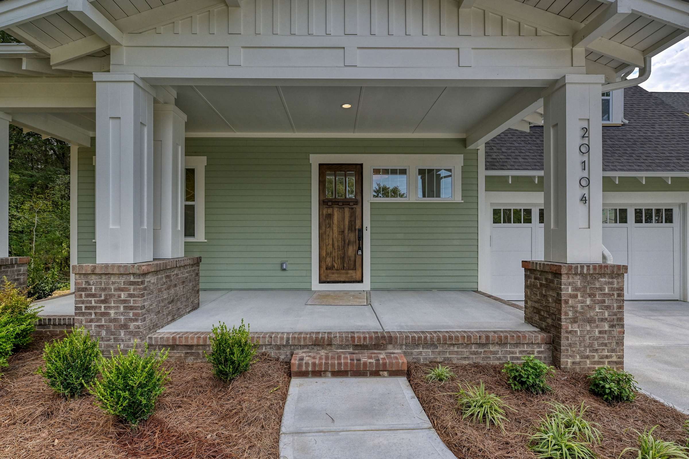 Covered front porch with brick piers, sage green siding, and a wood front door