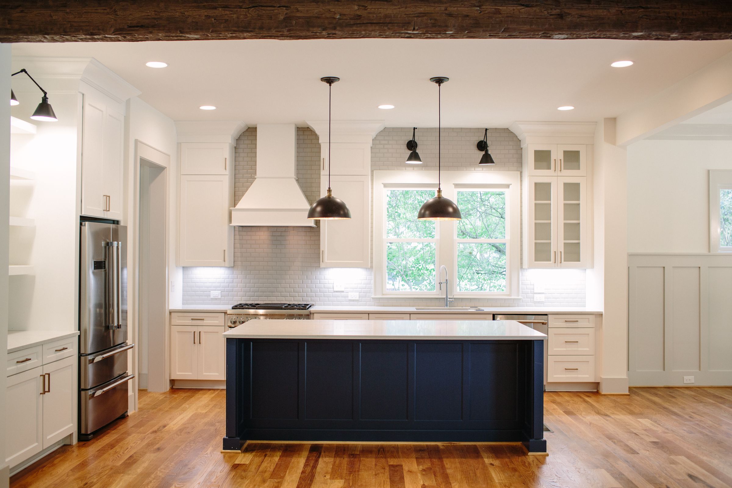 Kitchen with a navy island, white cabinets, a range hood, black pendants, and a window over the sink